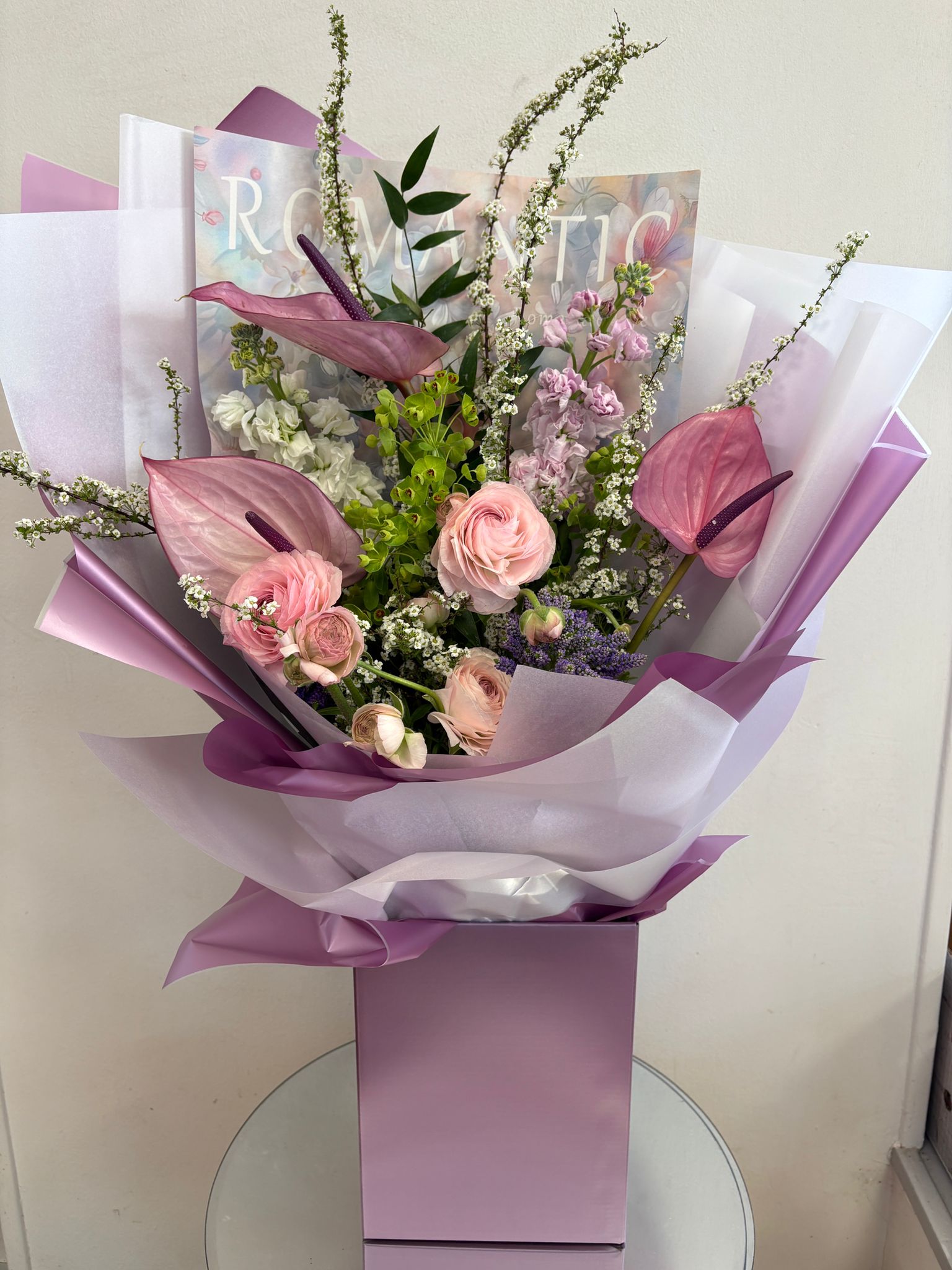 Hand-tied gift bouquet of ranunculus, roses and anthuriums in lilac wrap by Pure Flowers of Distinction